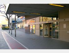 Eingangsbereich eines modernen Gebäudes mit Glasfronten, automatischen Türen und einem Vordach. Im Hintergrund sind weitere Gebäude, ein Fahrradweg sowie eine Bushaltestelle zu sehen.