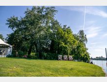 Grüne Wiese mit mehreren hohen Bäumen am Ufer eines Flusses, auf dem Wasser sind Industrieanlagen in der Ferne sichtbar. Am rechten Bildrand stehen drei Strandkörbe, und links befindet sich ein kleines Gebäude mit Zeltüberdachung.