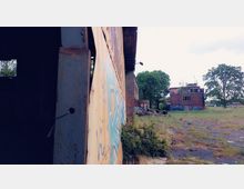 Verlassene Industrieanlage mit einer rostigen Metallwand im Vordergrund, bedeckt von Graffiti. Im Hintergrund sind ein rotes Backsteingebäude mit bunten Graffiti und überwachsene Vegetation sowie Bäume sichtbar.