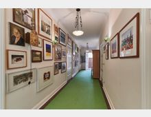 Langer, schmaler Flur mit hellem Anstrich, grünem Teppichboden und weißer Deckenbeleuchtung. Wände sind dicht mit gerahmten Bildern und Kunstwerken dekoriert, links hängt eine antike Wanduhr; am Ende des Flurs befindet sich eine geöffnete Tür, die einen Blick nach draußen ermöglicht.