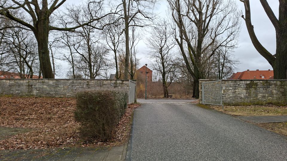 Einfahrt zu einem Park oder Friedhof mit asphaltierter Zufahrt zwischen niedrigen Steinmauern und einem geöffneten Metalltor. Dahinter stehen hohe, unbelaubte Bäume; im Hintergrund sind rote Ziegeldächer und ein Ziegelgebäude zu sehen.