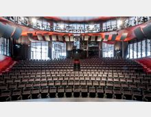 Moderner Theatersaal mit ansteigenden Sitzreihen aus schwarzem Stoff, roten Treppenstufen auf beiden Seiten und großen Glasfassaden im Hintergrund. Oben befinden sich Bühnenlichter und ein Technikbereich mit Vorhängen.