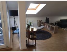 Wohnbereich in einer Dachgeschosswohnung mit Holzfußboden, Dachflächenfenster und moderner Einrichtung. Zu sehen sind ein Sessel auf einem runden Teppich, ein kleines Regal mit Büchern und Deko, ein Sofa im Hintergrund sowie eine Stehlampe und ein Glastürzugang zu einem Balkon links.