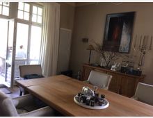 Esszimmer mit einem großen rechteckigen Holztisch und gepolsterten Stühlen. Im Hintergrund stehen ein Sideboard mit Dekorationen, ein großes Gemälde an der Wand, ein Kerzenleuchter und eine geöffnete Glastür mit Blick auf eine Terrasse.