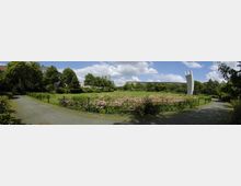 Weitläufige Parkanlage mit Rasenflächen und blühenden Blumenbeeten, umgeben von Bäumen und vereinzelten Gebäuden im Hintergrund. Rechts steht eine hohe, moderne Steinskulptur mit zwei gebogenen Säulen.