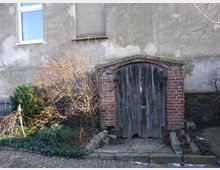 Alte, hölzerne Tür in einem gemauerten Rundbogen aus roten Ziegeln, neben Sträuchern und Pflanzen, vor einer verwitterten Hausfassade mit einem Fenster.