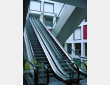 Breite Rolltreppe in einem modernen Innenraum mit weiß gestrichenen Wänden und großen, quadratischen Fenstern. Die Umgebung wirkt lichtdurchflutet, und farbige Akzente sind an den Wänden unterhalb der Fenster erkennbar.