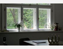 Ein heller Innenraum mit einem weißen, mehrteiligen Fenster, durch das grüne Bäume sichtbar sind. Auf der Fensterbank stehen eine weiße Vase mit Blumen, ein kleines Dekoobjekt und eine Bärenfigur, davor ein Tisch mit einem Kerzenständer.