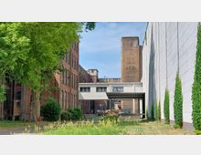 Industriegelände mit Backstein- und Betongebäuden, flankiert von rankenden Pflanzen und einem Baum auf der linken Seite. Im Hintergrund sind ein Verbindungsgang und ein hoher, verwitterter Betonturm zu sehen.