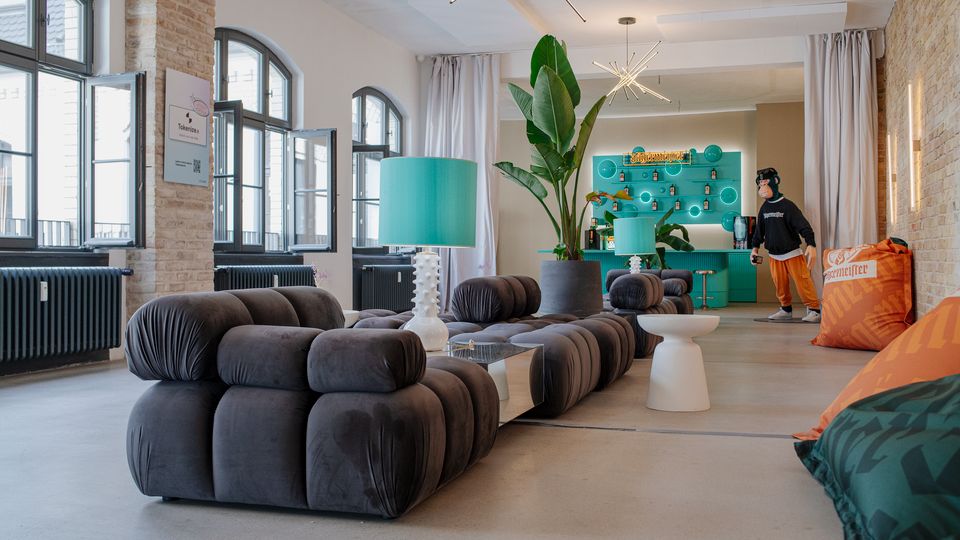 Heller Lounge- und Barbereich in einer umgebauten Industriehalle mit hohen Rundbogenfenstern, freigelegten Ziegelwänden und Betonboden. Große modulare Sofaelemente, türkisfarbene Leuchten, eine große Topfpflanze und eine türkisfarbene Bar mit beleuchteten Regalen prägen den Raum.