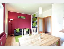 Wohnzimmer mit Holzboden und einer Wand in kräftigem Rot. Das Zimmer ist möbliert mit einem schwarzen Sofa, grünen Kissen, einem Holzesstisch mit Stühlen und einem Stehlampe in der Ecke; an der Wand hängen Naturbilder. Türen und dekorative Details wie ein Teppich und Blumen in einer Vase runden den Raum ab.