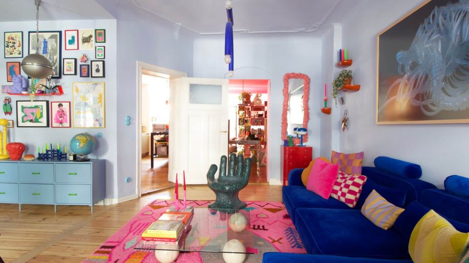 Bright, eclectic living room with pale blue walls, a deep blue sectional sofa with colorful pillows, and a pink patterned rug around a glass coffee table. The space features a gallery wall of framed art above a low cabinet, a sculptural hand-shaped chair, hanging pendant lights, and double doors opening to an adjoining room.
