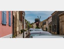 Eine schmale, gepflasterte Straße mit parkenden Autos, gesäumt von farbigen Häusern mit traditionellen Fassaden und blauen Fensterläden; im Hintergrund steht eine historische Windmühle unter blauem Himmel.