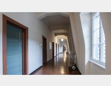 Ein langer, schmaler Flur mit poliertem braunem Boden, weißen Wänden und einer Strukturdecke mit sichtbaren Balken. Auf der linken Seite befinden sich Türen mit braunen Rahmen, rechts ein großes Fenster, das Tageslicht hereinlässt; im Hintergrund steht ein Rollwagen.