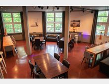 Ein moderner Raum mit roten Bodenfliesen, mehreren Tischen und Stühlen, großen Fenstern mit Blick ins Grüne und schlichter Dekoration, darunter gerahmte Bilder an den Wänden. Der Raum wird durch Tageslicht und Deckenleuchten beleuchtet, mit einem Mix aus Sitzbereichen und langen Tischen.