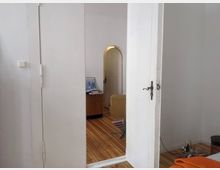 Innenaufnahme einer Wohnung mit offenem Durchgang. Zu sehen sind weiße Wände, eine geöffnete Zimmertür, Holzdielenboden, ein weiterer Raum mit einem gelben Sessel und Möbeln sowie kleinere Gegenstände im Vordergrund.