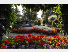 Gepflegter Garten mit bunten Blumenbeeten in Rot, Lila, Gelb und Orange, eingerahmt von grünen Sträuchern und Bäumen. Im Hintergrund befindet sich ein dekorativer Springbrunnen, umgeben von symmetrisch angelegten Wegen und Laubbögen.