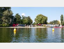 Seeufer mit grünem Wasser, gelben Bojen und einem kleinen Sandstrand. Im Hintergrund stehen Bäume, ein Weidenbaum, Sitzplätze mit Sonnenschirmen und Gebäude, darunter ein Pavillon sowie ein Bereich mit roten Zelten.