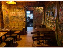 Schmaler Innenraum einer Bar oder eines Cafés mit Holzboden, warmem Deckenlicht und dicht mit Graffiti und Stickern bedeckten Wänden. Links stehen ein Sofa und kleine Tische, rechts mehrere Klapptische mit Stühlen; im Hintergrund führt ein Durchgang zu einem helleren Bereich.