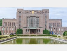 Backstein-Gebäude im symmetrischen Stil mit großen Fensterflächen und einem goldfarbenen Symbol an der Fassade. Im Vordergrund befindet sich ein rechteckiger Teich, umgeben von Grünflächen und Sträuchern.
