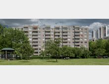 Mehrstöckiges Wohngebäude aus Betonplattenbau in einer grünen Parkanlage mit Rasenfläche, vereinzelten Bäumen und einem kleinen Pavillon. Im Hintergrund sind weitere Hochhäuser und dichter Baumbewuchs zu sehen, während dunkle Wolken den Himmel bedecken.