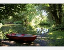 Rotes Ruderboot am Ufer eines kleinen Sees, umgeben von dichtem Wald mit grünem Laub, das sich im ruhigen Wasser spiegelt. Die Szene ist im Schatten und von Sonnenlicht durchbrochen.