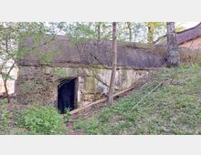 Eine alte, überwucherte Backsteinstruktur mit flachem Dach und einem offenen Eingang, gelegen in einem bewaldeten Gebiet. Im Inneren des Eingangs ist eine Person erkennbar, während die Umgebung von Gras, Bäumen und Sträuchern dominiert wird.