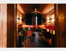 Blick in einen schmalen Bar- oder Loungebereich mit freigelegten Backsteinwänden, roter gepolsterter Sitzbank und kleinen runden Tischen mit Stühlen. An der Decke hängt ein Ventilator, am Ende des Raums sind dunkle Vorhänge, seitliche Wandleuchten und ein Holzrahmen am Eingang zu sehen.