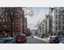 Straßenszene in einem Wohnviertel mit Altbauten: Links rötliche Backsteingebäude, rechts hell verputzte Häuser mit Balkonen. Geparkte Autos säumen die Straße, darüber kahle Bäume und ein bewölkter Himmel.