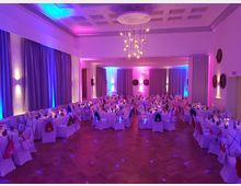 Ein festlich dekorierter Veranstaltungssaal mit gedeckten runden Tischen und weißen Stuhlhussen, beleuchtet durch farbige Lichter in Blau, Rosa und Violett. Die Decke ist mit modernen Hängeleuchten ausgestattet, und bodenlange Vorhänge bedecken die Fenster an den Wänden.