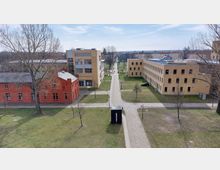 Blick auf einen Campus mit grünen Rasenflächen, Bäumen und gepflasterten Wegen. Umgeben von Gebäuden in rotem Ziegel und gelblichem Putz, mit einem kleinen modernen Pavillon in der Mitte.