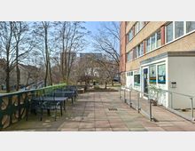 Terrasse vor einem mehrstöckigen Gebäude mit großen Fenstern, einem Eingang mit Glastür und einer Rampe mit Geländer. Links stehen mehrere Metalltische und -stühle entlang einer grünen Brüstung; im Hintergrund sind Bäume, Wohnhäuser und eine Straße zu sehen.