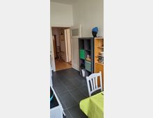 Schmale Küche mit dunklem Fliesenboden und heller Wandgestaltung. Zu sehen sind ein offenes Regal mit Haushaltsgegenständen, ein gedeckter Tisch mit grünem Tischtuch, und ein Durchgang zu einem angrenzenden Raum mit Holzboden.