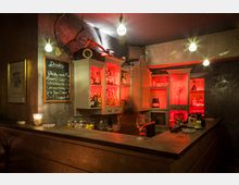 Gemütliche Bar mit rustikalem Tresen, roten Lichteffekten und Holzschränken, die mit Flaschen und Gläsern gefüllt sind. Eine Tafel mit handgeschriebenem Getränkemenü hängt an der Wand, Dekorationen wie Geweihe und Kerzen ergänzen das Ambiente.