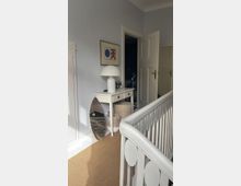 Ein helles Inneneck mit einem weißen Tisch, darauf eine weiße Tischlampe und ein gerahmtes Bild an der Wand. Daneben stehen ein gestreiftes Brett und zwei geflochtene Körbe, der Boden ist mit einem beigefarbenen Teppich bedeckt, und eine weiße Holzgeländerbalustrade ist im Vordergrund sichtbar.