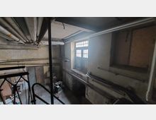 Ein altes, verlassenes Kellerraum mit sichtbaren Rohren an der Decke, verwitterten Wänden und einem großen Fenster mit Gitterstruktur. Im Raum befinden sich verschiedene Metall- und Holzobjekte sowie alte Maschinen.