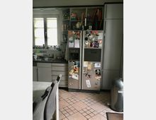 Eine moderne Küche mit grauen Schränken und einer doppelten Edelstahl-Kühlschranktür, die mit Magneten und Fotos dekoriert ist. Links befindet sich ein Fenster mit Jalousien, Spüle und Arbeitsfläche; rechts ein silberner Mülleimer auf einem gefliesten Boden.