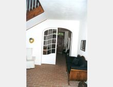 Innenraum eines Hauses mit hell gestrichenen Wänden und einem Boden aus roten Ziegelsteinen. Eine hölzerne Treppe führt nach oben, daneben steht ein gepolstertes Holzsofa, und durch eine Glastür mit geschwungenem Rahmen ist ein Flur mit hölzerner Eingangstür sichtbar.