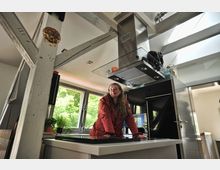 Moderne Küche mit zentralem Kochfeld und Dunstabzugshaube, umgeben von großen Fenstern, die Tageslicht hereinlassen. Sichtbare Holzbalken verstärken den rustikalen Charme, während eine Person an der Kücheninsel steht.