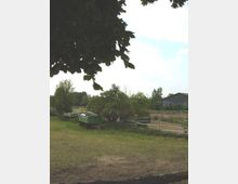 Eine ländliche Szene mit offenen Wiesen, mehreren grünen Anhängern und einem umzäunten Areal im Hintergrund. Im rechten Bildbereich ist eine große Scheune zu sehen, umgeben von Bäumen und Vegetation.