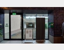 Blick durch eine gläserne Eingangstür mit Notausgangsschild in einem Gebäude. Im Vordergrund befinden sich eine Infotafel und ein Defibrillator an der Wand, im Hintergrund ist ein überdachter Tunnel zu sehen, durch den Tageslicht einfällt.