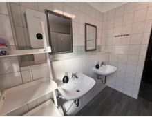 Gefliester Waschraum mit zwei wandmontierten Waschbecken, Seifenspendern und zwei Spiegeln über einer langen Ablage. Links steht ein offenes Regal mit einem wandhängenden Warmwassergerät; der Boden ist dunkel und wirkt wie Vinyl oder Laminat.