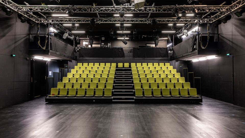 Innenraum eines schwarzen Theater- oder Studiorsaals mit ansteigender Tribüne aus hellgrünen Sitzreihen und mittigem Treppenaufgang. Über der Bühne hängen Metalltraversen mit Scheinwerfern und Technik, vor der Tribüne liegt eine freie, dunkle Spielfläche.