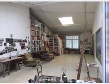 Ein geräumiges Künstleratelier mit hohen Decken und weiß gestrichenen Wänden. Der Raum enthält Regale mit Büchern und Materialien, eine Collage aus Fotos an der Wand, Malutensilien auf Tischen und mehrere Sitzgelegenheiten; ein Oberlicht und ein großes Fenster lassen Tageslicht herein.