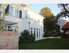 Zweistöckiges modernes Wohnhaus mit weißer Fassade und Balkontüren im Erdgeschoss, umgeben von einem Garten mit Rasen, Bäumen und einem kleinen Holzgerüst links. Im Hintergrund befindet sich ein Nebengebäude mit rotem Ziegeldach.