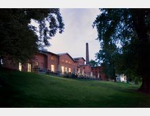 Außenansicht eines historischen Backsteingebäudes mit großen Fenstern und einem Schornstein, umgeben von Bäumen und einer Rasenfläche, aufgenommen bei Dämmerung. Beleuchtete Fenster und außenliegende Sitzbereiche lassen auf eine Gastronomienutzung schließen.