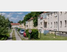 Wohnsiedlung mit zweistöckigen, beigen Mehrfamilienhäusern, die entlang einer schmalen Straße mit parkenden Autos stehen. Auf der rechten Seite befindet sich eine Wiese mit einem Wäscheständer und aufgehängter Wäsche; im Hintergrund Bäume und blauer Himmel.