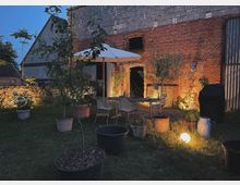 Hof bei Dämmerung mit Sitzbereich unter einem weißen Sonnenschirm, umgeben von Topfpflanzen und einer beleuchteten Ziegelsteinfassade eines alten Gebäudes. Im Vordergrund sind Grasflächen und mehrere Pflanzkübel zu sehen, die Szene wird von warmem Licht akzentuiert.