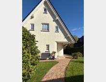 Wohnhaus mit heller Putzfassade und steilem Satteldach, eingerahmt von einem gepflegten Garten mit Rasenflächen und Pflanzen. Ein gepflasterter Weg führt zum Eingangsbereich, der mit kleinen Stufen und Blumen in Töpfen dekoriert ist.