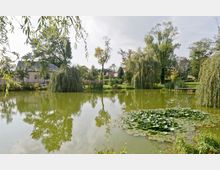 Ein idyllischer Teich mit grünem Wasser, umgeben von Weidenbäumen und anderen Bäumen. Im Hintergrund stehen ein Haus mit hellen Wänden und einem Schieferdach sowie weitere Gebäude mit roten Dächern, eingebettet in eine parkähnliche Landschaft.